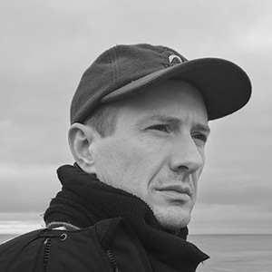 Black and white close-up of a man wearing a cap and scarf, looking off to the side outdoors.