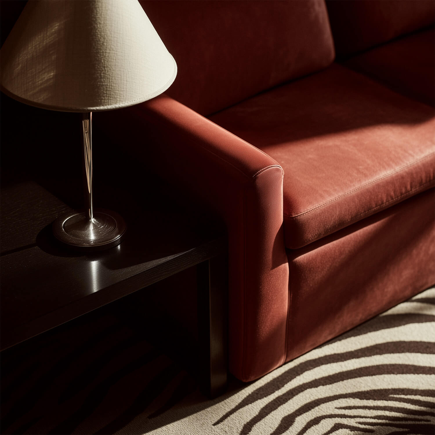 Close-up of a red sofa arm next to a dark wooden side table with a white lampshade.