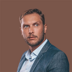 Portrait of a man with short hair and beard, wearing a blue blazer and white shirt, against a brown background.