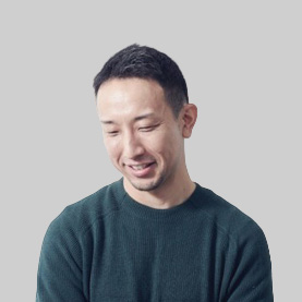 Smiling man with short black hair in a dark green sweater looking down against a gray background.