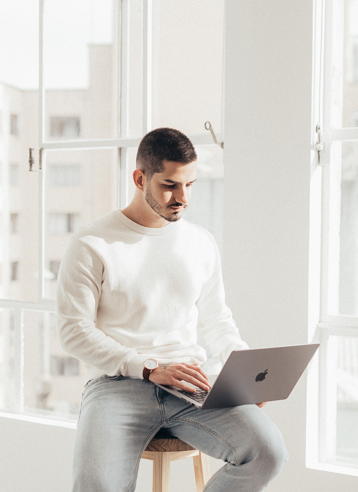 Man wearing white sweater and jeans sitting on stool using a MacBook laptop near large windows.