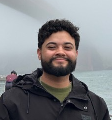 Smiling man with beard and curly hair wearing a black jacket near water on a foggy day.