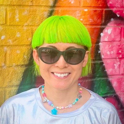 Person with neon green hair, black sunglasses, smiling, wearing a colorful beaded necklace and silver shirt.